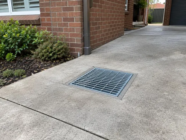 Outdoor drain grate set into dry concrete paving beside a brick house in Westmead