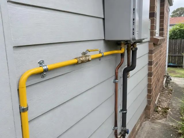 Yellow gas piping and meter on an exterior weatherboard wall beside a brick side passage in Parramatta